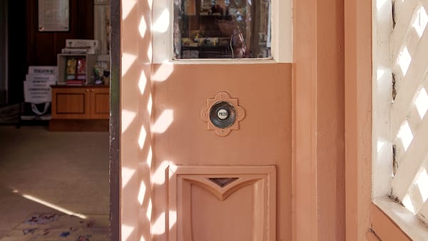 An open front door of a house at midday with sunlight falling across a woven mat and a pair of boots on the step