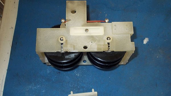 A kitchen wall clock lying face up on a table with its back panel removed, a small screwdriver resting beside the loose hour hand.
