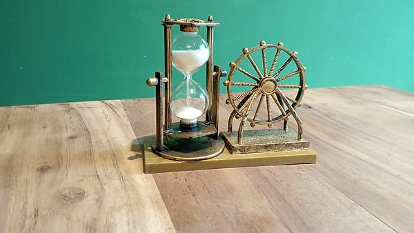 A mechanical stopwatch lying face up on a scratched wooden desk beside a folded paper notebook
