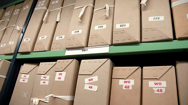 An open cardboard storage box on a concrete floor with loose printed pages stacked unevenly inside it