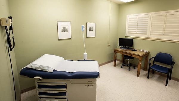 A small consulting room with two chairs at an angle, a clipboard on the seat of one and a boxed tissue on the sill