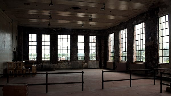 A tall paneled room with rows of empty wooden seats facing a raised platform beneath high windows
