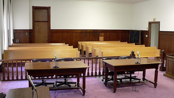 An empty lectern facing a raised bench in a quiet room with pale walls and a row of tall windows