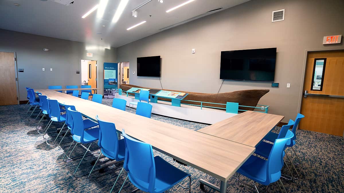 A narrow shaft of daylight falling across an empty conference table and two stacked chairs in a bare room