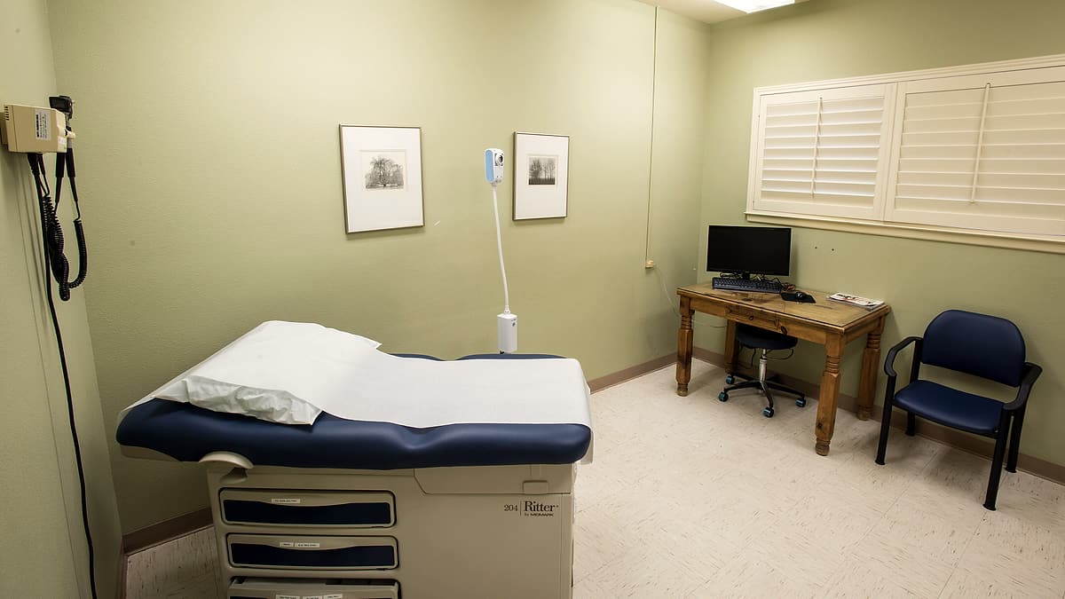A small consulting room with two chairs at an angle, a clipboard on the seat of one and a boxed tissue on the sill
