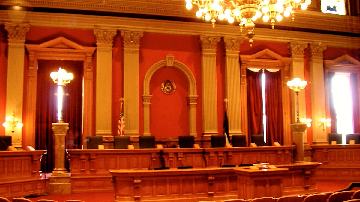 A wooden courtroom bench with a closed binder, a microphone on a short stand and an empty water glass beside it